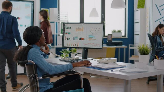Invalid disabled handicapped african businesswoman sitting in wheelchair working at computer with financial statistics talking at phone with partners in start up business office, modern workspace