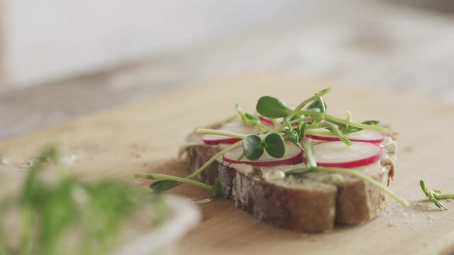 The man is cooking in the kitchen. Man with microgreens in hand sprinkles radish sandwich. Making a vegan sandwich step by step. Slice of rye bread with radish and microgreens on a wooden background