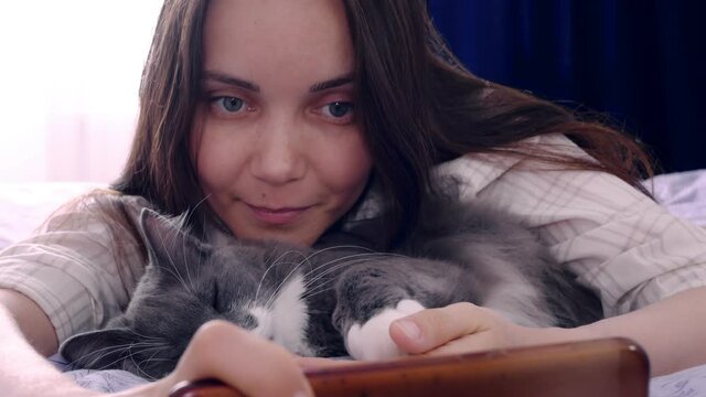 A young woman lies on a bed with a fluffy beautiful cat. The cat has a white tie around its neck and soft white paw pads. Cute woman takes selfie pictures on a smartphone. Love for domestic cats.