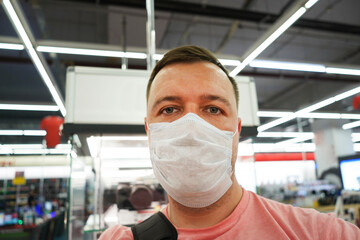Fototapeta na wymiar young man in medical mask looks at camera in electronics store against background of display case with cameras. new normal during trip to shop