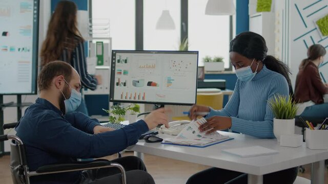 Invalid businessman and african employee with protective mask against coronavirus working together at financial project in new normal start up company, comparing statistics looking at computer