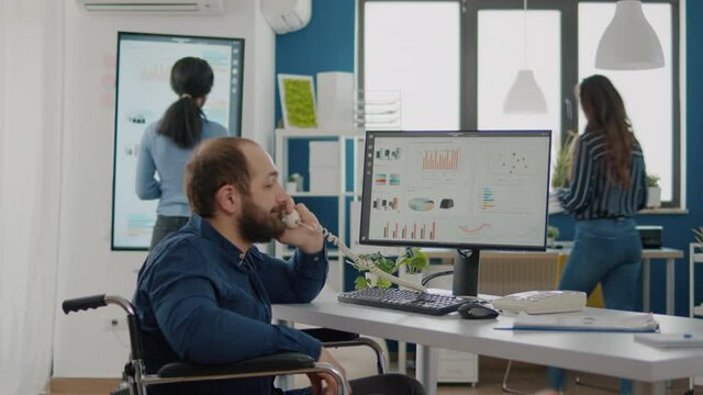 Invalid disabled handicapped businessman sitting in wheelchair working at computer with financial statistics answering at phone talking with partners in start up business office, modern workspace