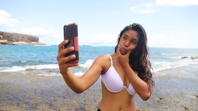 Young Latina girl takes selfies on the beach and sticks out her tongue making grimaces and funny faces.
