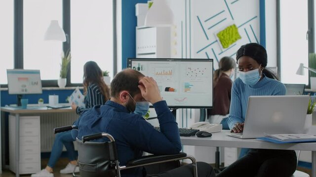 Diverse coworkers with protection face masks working together in new normal workplace during pandemic, invalid manager with disabilities pointing at laptop explaning to african woman company startegy