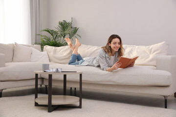 Young woman reading book on sofa at home - Powered by Adobe