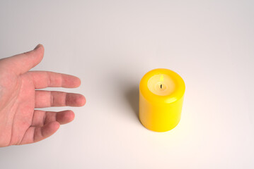 A hand reaches for a yellow lighted candle on a white background.