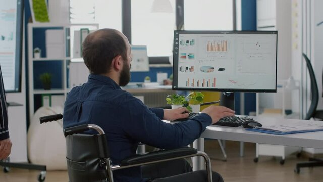 Immobilized paralysed handicapped manager analysing graphs and typing on computer sitting in wheelchair in start up business office, while diverse team working at financial project in background.