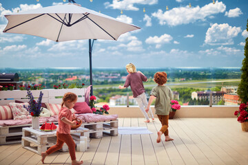 cheerful kids having fun, running on rooftop patio with pallet lounge space, umbrella and decking,...