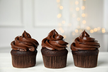 Delicious chocolate cupcakes with cream on white wooden table against blurred lights. Space for text