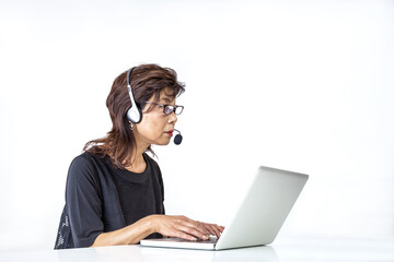 白背景でヘッドセットを付けノートパソコンを操作するシニア女性
