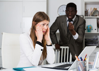 Upset business woman sitting at laptop in office on background with angry colleague