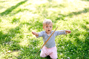 Small child is kneeling on a green lawn and holding a twig in his hands