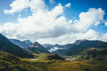 Scenic green blue alpine landscape with mountain lake in highland valley in sunlight and big glacier under cloudy sky. Shadow of clouds on green mountain valley. Clouds shadow on rocks and hills.