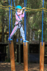 Fototapeta premium The child is engaged in sports.A girl overcomes sports obstacles in a rope park.A little girl in a protective helmet and tracksuit.Active recreation in the summer.Sports Rope Park for children