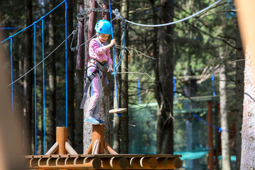 The child is engaged in sports.A girl overcomes sports obstacles in a rope park.A little girl in a protective helmet and tracksuit.Active recreation in the summer.Sports Rope Park for children