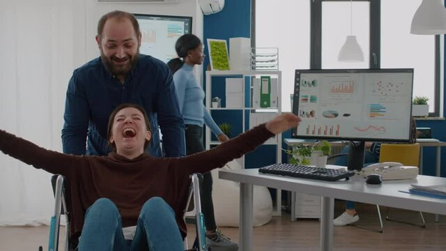 Cheerful employee taking break pushing wheelchair with his paralysed invalid disabled colleague smiling and having fun in start up financial business office. Immobilized woman laughing in workplace