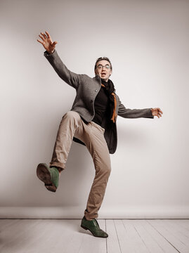 Adult man hipster in pants and plaid jacket stands holding foot up, making giant step, is out of balanced, falling down waving hands over grey background