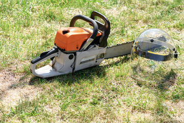 Naklejka premium Chainsaw and protective face shield, helmet on the green grass on a sunny day. Concept is to cut trees