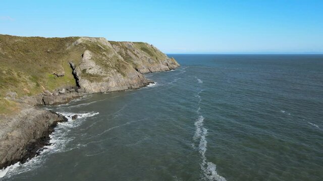 Waves Hitting Limestone Cliffs Near Three Cliffs Bay With Blue Sky In The Background In Swansea, Wales. - aerial