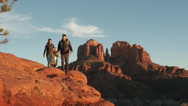 A caucasian family of three with outerwear hiking red sandstone cliffs and admiring the view with a clear blue sky at sunset near Sedona, Arizona.