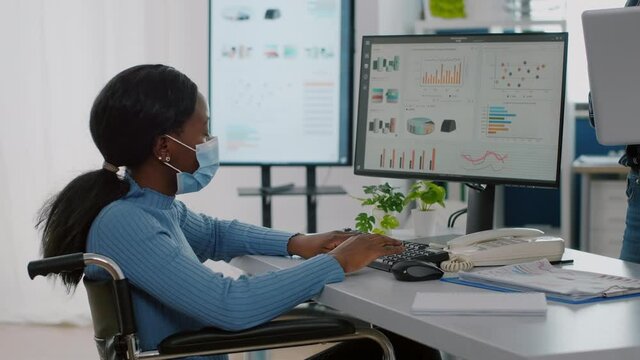 Handicapped paralysed invalid african employee with medical face mask discussing with coworker economic project pesenting strategy of company. Multiethnical team working in new normal startup office