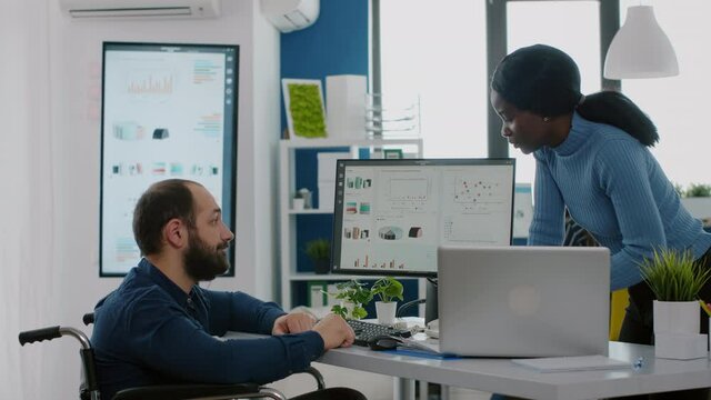 Invalid disabled paralysed bookkeeper in wheelchair working with african woman colleague calculating profit on laptop, taking notes on pc, analysing financial documents in start-up financial company