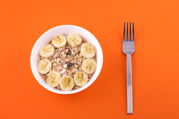 oatmeal in white plate with sliced banana and fork on orange background, healthy food concept