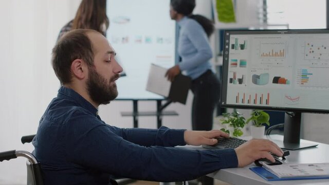 Paralysed immobilized invalid worker with disabilities working at computer analysing financial statistics company efficiency typing on pc sitting in wheelchair. Diverse team discussing in background