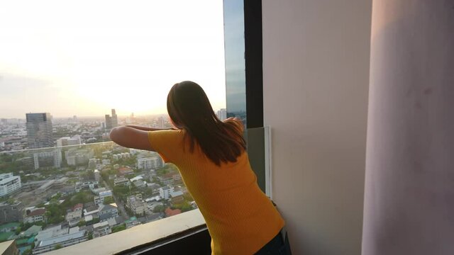 Beautiful Asian woman standing on apartment balcony looking away to cityscape and skyscraper at sunset with happiness. Smiling female relax and enjoy city life and indoor lifestyle at condominium