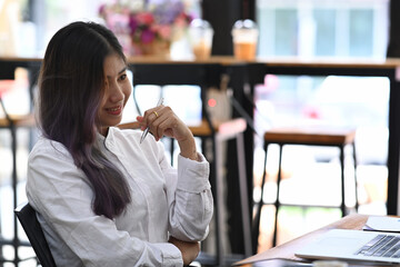 Smiling young woman holding pen and looking at laptop screen while sitting in office.