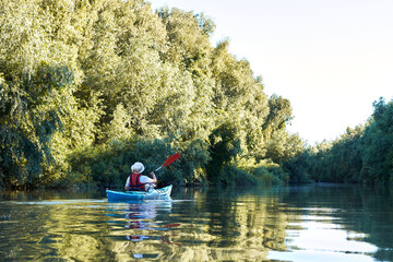 Fototapeta na wymiar Woman row in blue kayak at wilderness areas of Danube river near green trees and thickets of wild grapes at spring