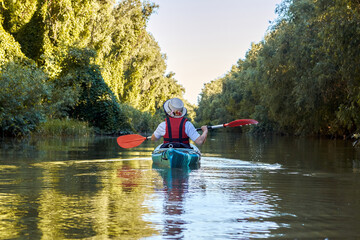 Fototapeta na wymiar Back view on woman row in blue kayak at wilderness areas of Danube river near green trees and thickets of wild grapes at spring