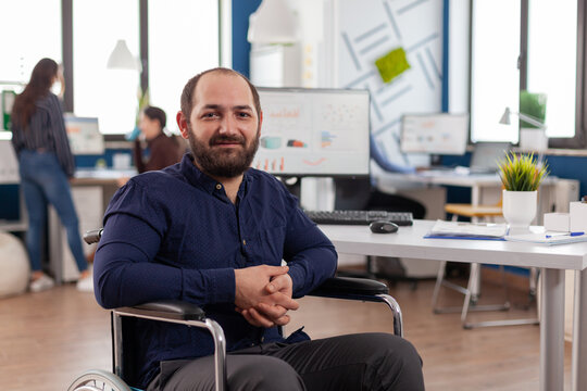 Project leder with disabilities, invalid handicapped disabled paralized looking at camera sitting immobilized in wheelchair in business office room working at financial project with team.