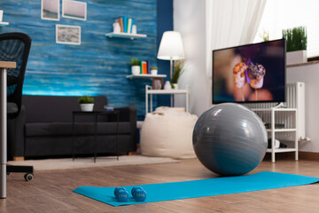 Empty living room with nobody in it ready for fitness training with yoga mat and dumbells on clean floor ready for training body myscle during wellness workout. Front view of sports equipment
