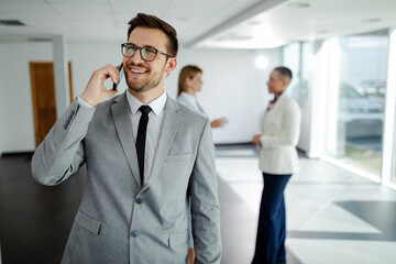 Young smiling businessman having phone call while standing in hallway before meeting.