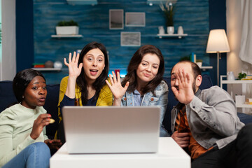 Happy multi-ethnic friends greeting her collegue during online videocall meeting using laptop webcam. Group of multiracial people spending time together on couch late at night during party