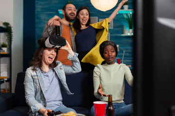 Happy young woman celebrating victory while playing video games with virtual reality headset and her multi ethnic friends are cheering up for her while socialising in the evening.
