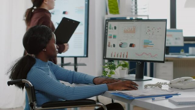 African disabled invalid manager woman working at pc showing data prosessing on desktop for project regarding economy looking at financial documents checking graphs, reading raports in business office