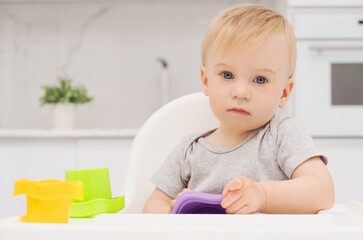 Cute caucasian sad baby girl sitting at high chair in kitchen, modern light interior, playing with toys, looking at camera. Close-up shot
