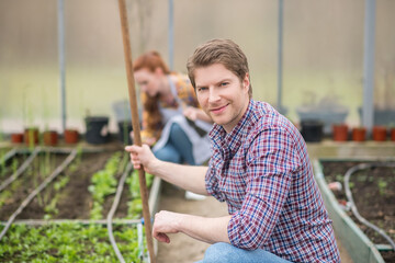 Friendly man with rake crouched near seedlings in greenhouse