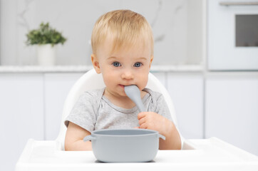 Adorable funny caucasian blonde baby girl eating from silicone plate at highchair in kitchen, with spoon in mouth, looking at camera. Healthy food for kid concept