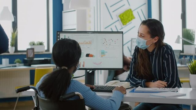 Freelancers wearing protective face masks working on computer in business office during global pandemic, disabled african woman sitting in wheelchair . Team discussing in new normal financial office