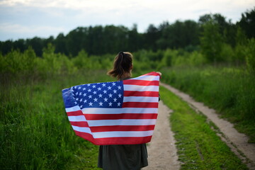 A young, happy American woman stands with her back turned, holding the US national flag fluttering in the wind on her shoulders, relaxing in the open air.