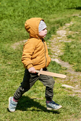 A child in a jacket and a hat with a wooden sword in his hands plays outdoors - 438365367