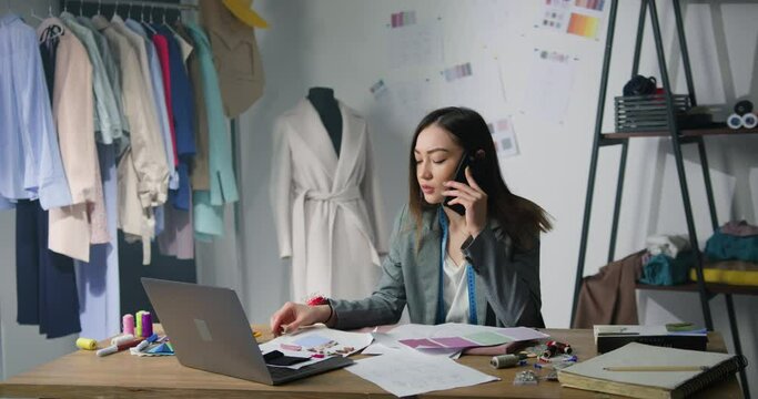 Nervous disappointed asian woman fashion designer sitting at atelier workplace looking at fabric samples and having unpleasant phone conversation. Job stress concept.
