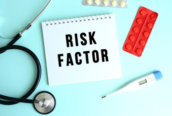 The text RISK FACTOR is written on a white notepad that lies next to the stethoscope and pills on a...