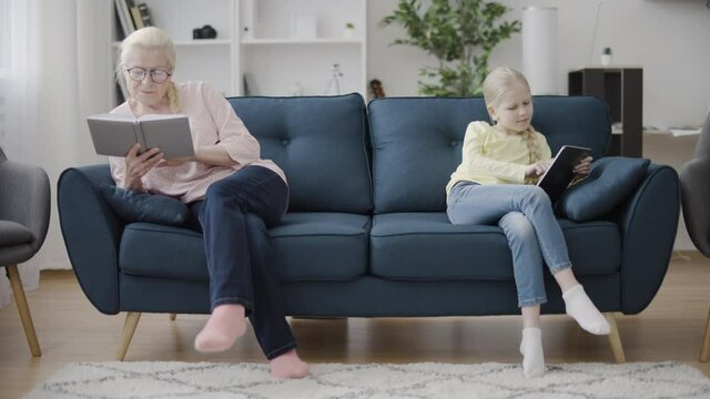 Granny reading book while little girl scrolling tablet, generation gap, family