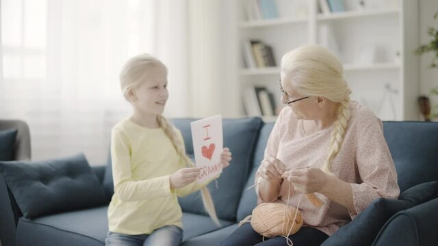 Cute girl presenting greeting card to granny knitting on sofa, happy family