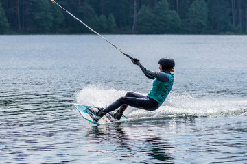 Girl with helmet and life vest rides a wakeboard on the lake with forest on background, wakeboarding, water sport in summer 