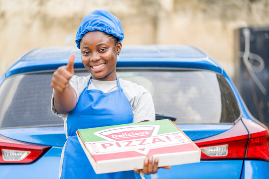 image of cheerful african lady in apron and head wrap behind automobile, carrying pizza with thumb up sign- pizza delivery concept